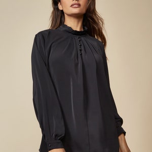 Puede incluir: Una blusa negra de manga larga con cuello alto y detalle de volantes. La blusa tiene cierre de bot&oacute;n en el cuello y un detalle fruncido en la parte delantera.