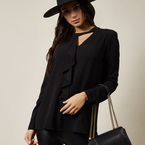 Pode incluir: Uma blusa preta de manga comprida com decote de babados e um detalhe de olho de fechadura. A blusa &eacute; combinada com leggings pretos e uma bolsa de m&atilde;o de couro preto com uma al&ccedil;a de corrente dourada.
