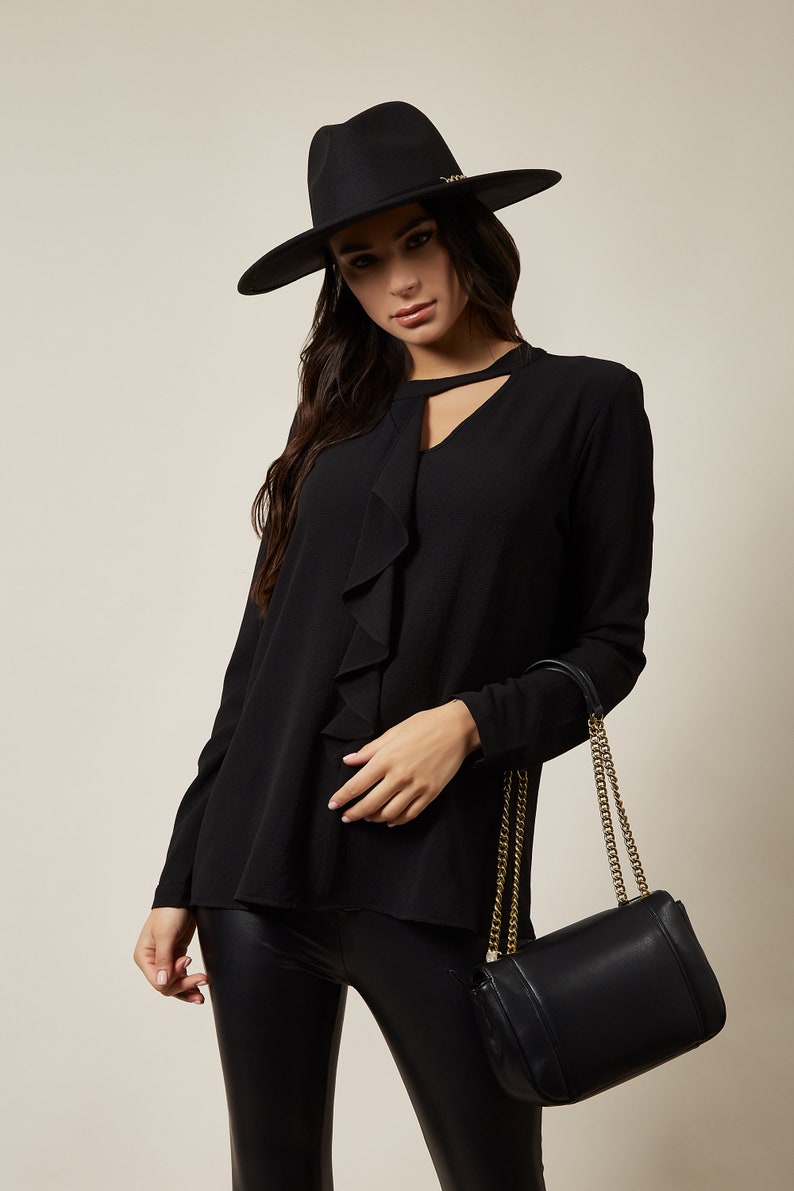 Pode incluir: Uma blusa preta de manga comprida com decote de babados e um detalhe de olho de fechadura. A blusa &eacute; combinada com leggings pretos e uma bolsa de m&atilde;o de couro preto com uma al&ccedil;a de corrente dourada.
