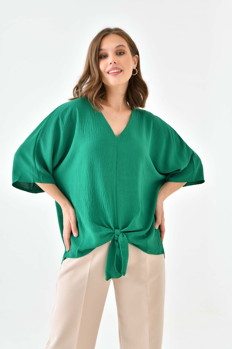 May include: A green, loose-fitting, v-neck top with a knotted front. The top has short, wide sleeves and is made of a textured fabric.