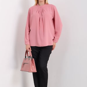 K&ouml;nnte beinhalten: Eine rosa Bluse mit langen &Auml;rmeln, einem R&uuml;schenkragen und Knopfdetails. Die Bluse ist in eine schwarze Hose gesteckt und die Person h&auml;lt eine rosa Handtasche.