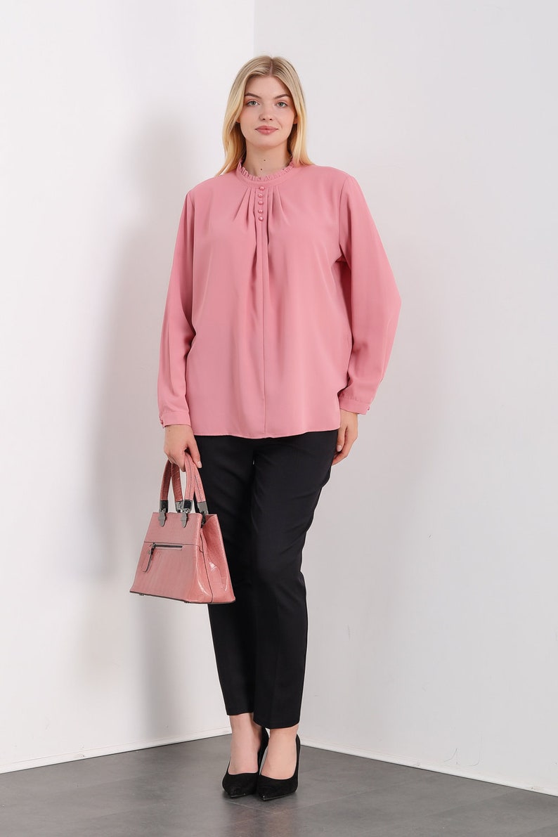K&ouml;nnte beinhalten: Eine rosa Bluse mit langen &Auml;rmeln, einem R&uuml;schenkragen und Knopfdetails. Die Bluse ist in eine schwarze Hose gesteckt und die Person h&auml;lt eine rosa Handtasche.