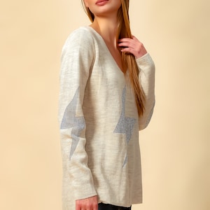 May include: A light beige sweater with a V-neck and silver glitter star designs on the sleeves.