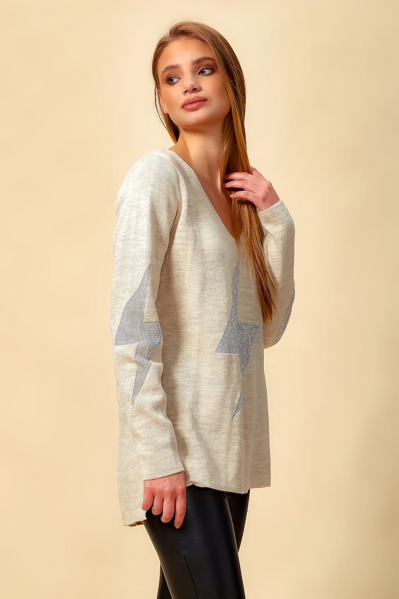 May include: A light beige sweater with a V-neck and silver glitter star designs on the sleeves.
