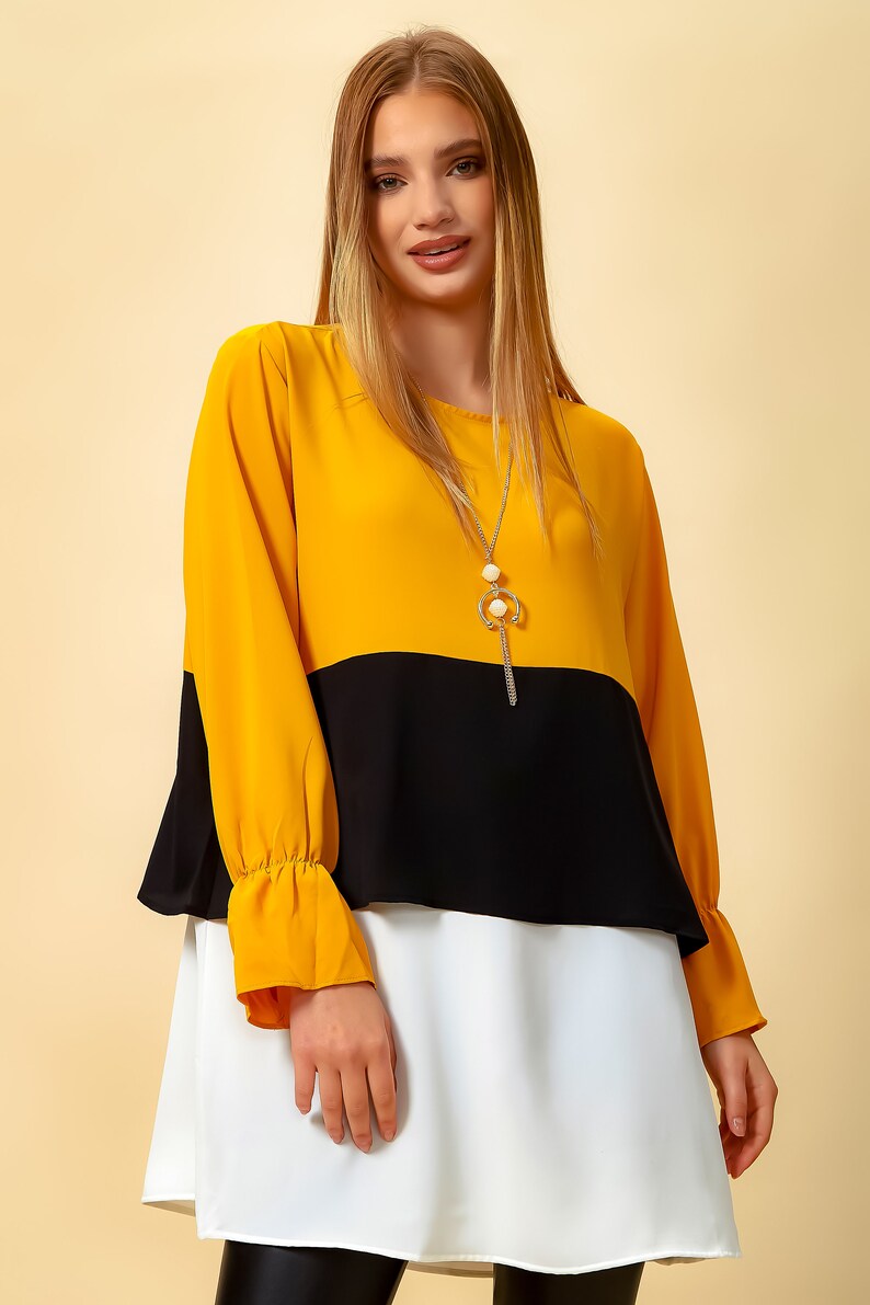 May include: A yellow, black and white layered tunic top with long sleeves and a gold chain necklace.