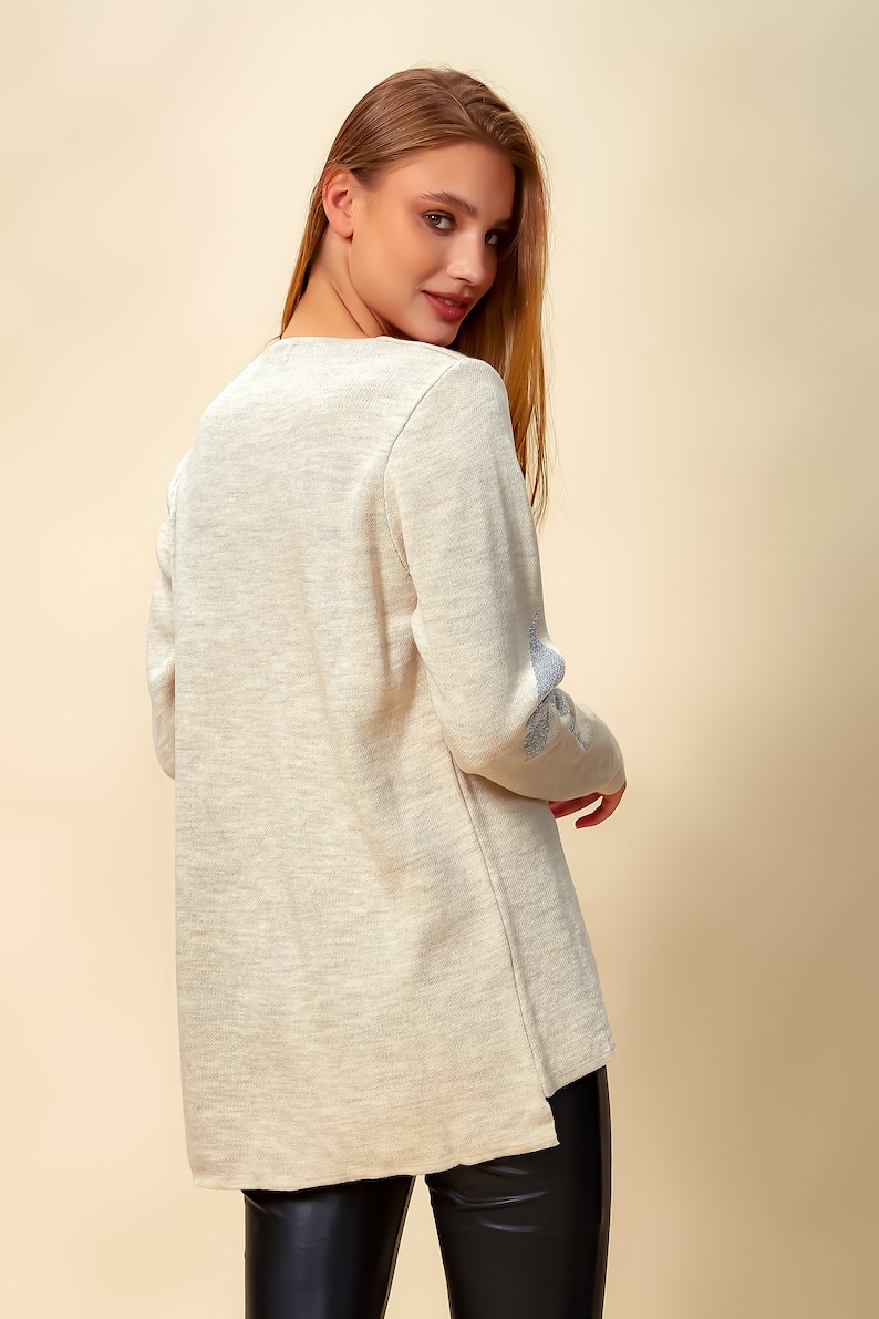 May include: A light beige long-sleeved sweater with a subtle texture and a blue embroidered design on the elbow.
