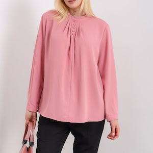 K&ouml;nnte beinhalten: Eine rosa Bluse mit langen &Auml;rmeln, einem R&uuml;schenkragen und einer Reihe von Kn&ouml;pfen vorne. Die Bluse ist aus einem leichten Stoff gefertigt und hat eine lockere Passform.