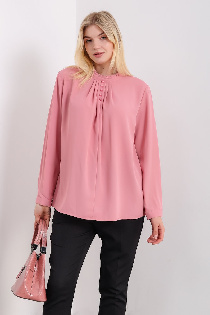 K&ouml;nnte beinhalten: Eine rosa Bluse mit langen &Auml;rmeln, einem R&uuml;schenkragen und einer Reihe von Kn&ouml;pfen vorne. Die Bluse ist aus einem leichten Stoff gefertigt und hat eine lockere Passform.