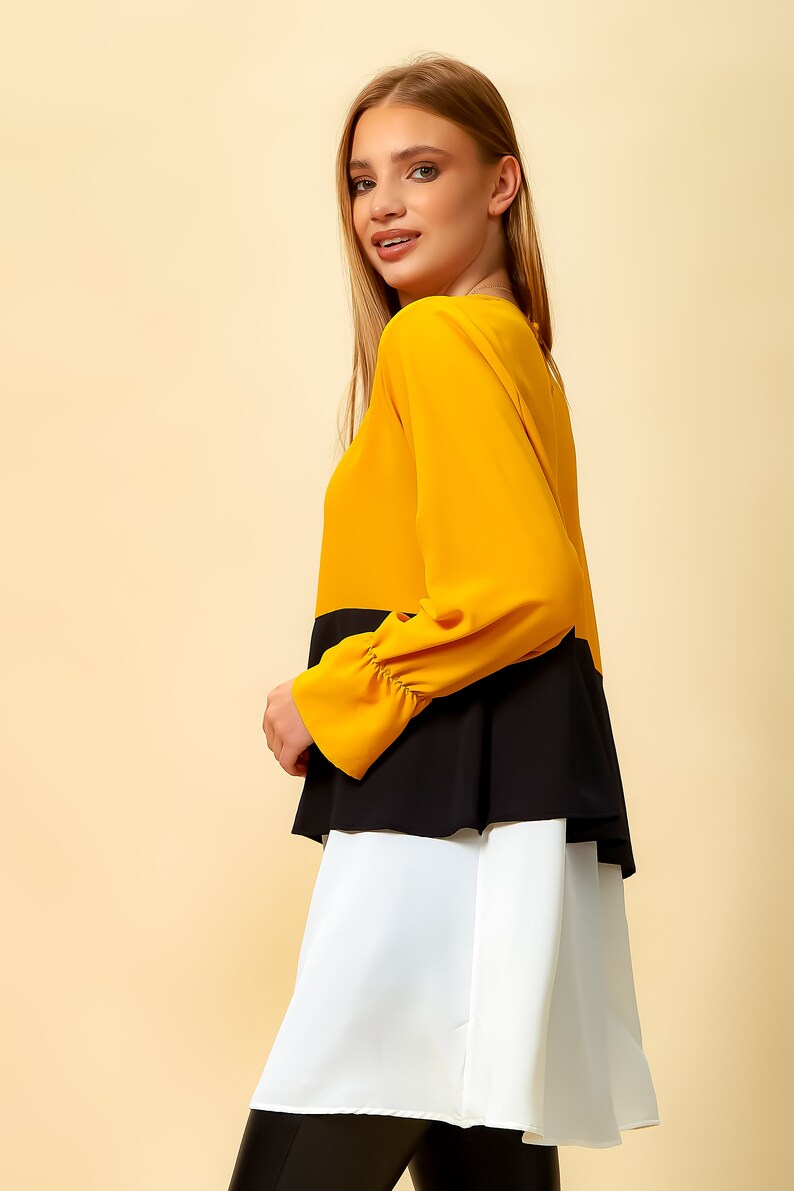 May include: A layered blouse with a mustard yellow top, black middle layer, and white bottom layer. The blouse has long sleeves with ruched cuffs.