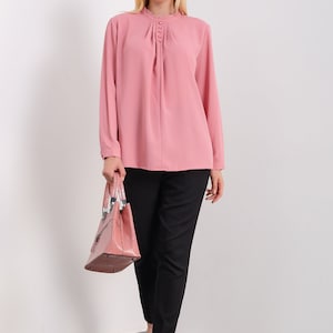 K&ouml;nnte beinhalten: Eine rosa Bluse mit langen &Auml;rmeln, einem R&uuml;schenkragen und Knopfdetails. Die Bluse ist in eine schwarze Hose gesteckt und die Person h&auml;lt eine rosa Handtasche.