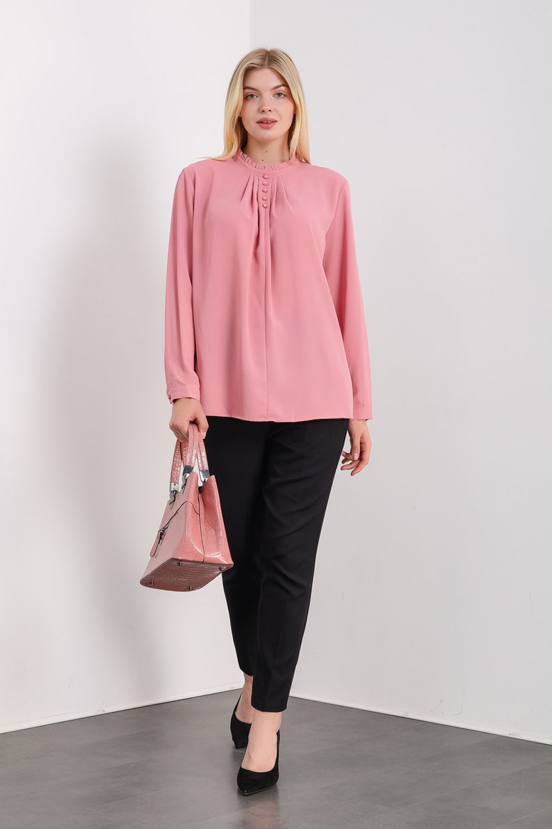 K&ouml;nnte beinhalten: Eine rosa Bluse mit langen &Auml;rmeln, einem R&uuml;schenkragen und Knopfdetails. Die Bluse ist in eine schwarze Hose gesteckt und die Person h&auml;lt eine rosa Handtasche.