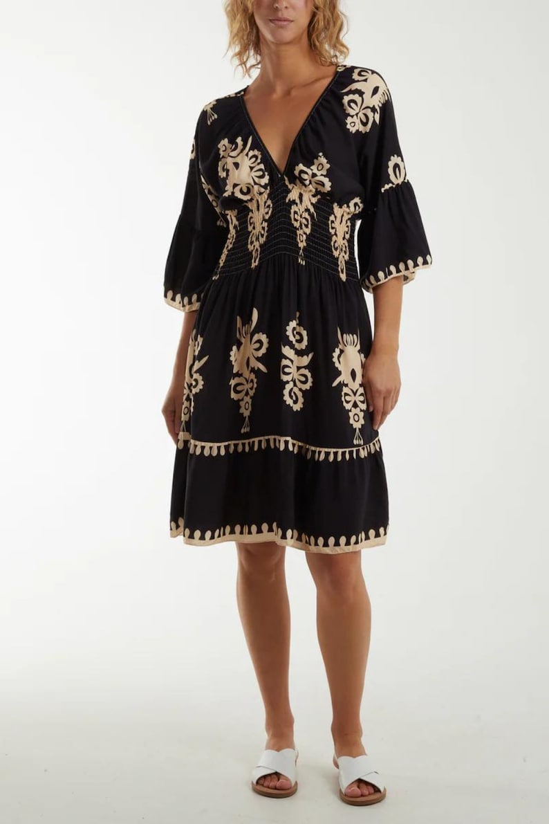 Puede incluir: Un vestido negro corto con cuello en V y mangas 3/4. El vestido presenta un estampado floral bordado en beige y una falda de capas. El vestido est&aacute; ajustado a la cintura con una banda el&aacute;stica.