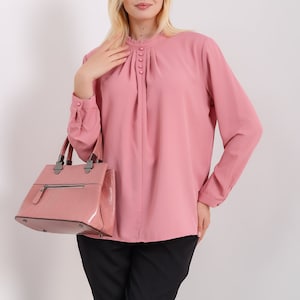 K&ouml;nnte beinhalten: Eine rosa Bluse mit langen &Auml;rmeln, einem R&uuml;schenkragen und Kn&ouml;pfen. Die Bluse hat eine gefaltete Vorderseite und wird mit einer schwarzen Hose getragen. Eine rosa Handtasche wird in der linken Hand getragen.