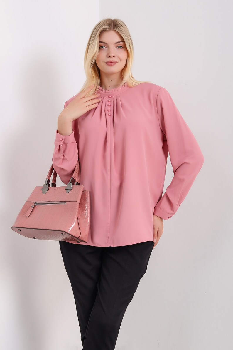 K&ouml;nnte beinhalten: Eine rosa Bluse mit langen &Auml;rmeln, einem R&uuml;schenkragen und Kn&ouml;pfen. Die Bluse hat eine gefaltete Vorderseite und wird mit einer schwarzen Hose getragen. Eine rosa Handtasche wird in der linken Hand getragen.