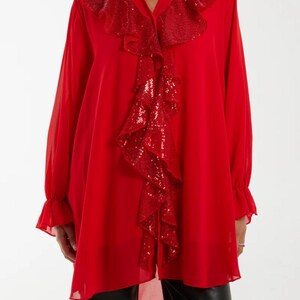 Puede incluir: Una blusa túnica roja vibrante con mangas largas y cuello en V. El escote y la parte delantera presentan un detalle de volantes con lentejuelas. Las mangas tienen puños con volantes. La blusa es de tejido ligero y fluido.