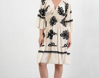 May include: Cream-colored dress with black floral embroidery and a V-neck. The dress features a smocked waist, three-quarter sleeves, and a tiered skirt. A straw hat and black sandals complete the outfit.