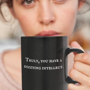 May include: Black coffee mug with the text "Truly, you have a dizzying intellect." The mug has a black handle and is held up in front of a person's face. The mug is a standard size.