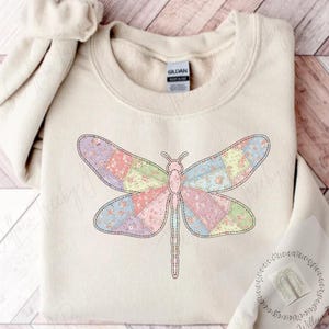 May include: Cream-colored sweatshirt featuring a colorful dragonfly design. The dragonfly's wings are composed of patchwork patterns in pastel shades of pink, blue, green, and yellow. The design is outlined with black stitching. The sweatshirt has a crew neck and long sleeves.