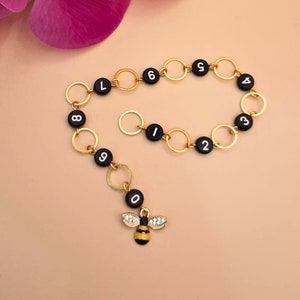 Könnte beinhalten: Ein goldenes Kettenarmband mit schwarzen Perlen, die die Zahlen von 0 bis 9 darstellen. Ein kleiner Bienenanhänger hängt am unteren Ende des Armbands.