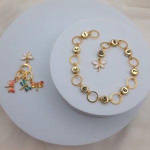 Könnte beinhalten: Ein goldenes Kettenarmband mit nummerierten Anhängern und einem Libellenanhänger. Ein separater Schlüsselanhänger mit drei Libellenanhängern und einem Schlüsselring.