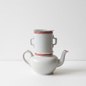 Könnte beinhalten: Eine weiße Keramik-Teekanne mit einer passenden Tasse, die obenauf gestapelt ist. Beide Teile haben rote und schwarze Verzierungen. Die Teekanne hat einen gebogenen Ausguss und Henkel. Die Tasse hat zwei kleine Griffe.