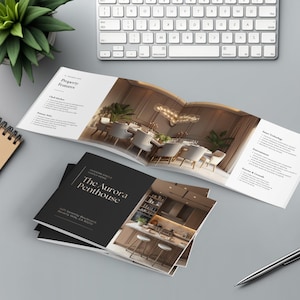 May include: An open brochure and a stack of brochures with the title "The Aurora Penthouse" on a gray surface. The open brochure displays a dining room scene. A keyboard, pen, coffee cup, and a plant are also visible.