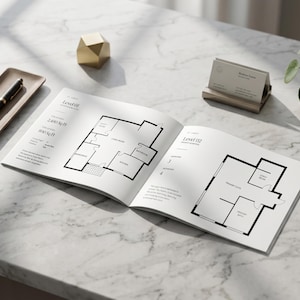 May include: An open architectural floor plan book with black line drawings of house layouts. A pen and tray, a gold geometric object, a business card holder, and a potted plant are on a marble surface.