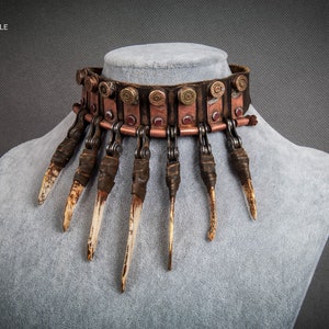 Puede incluir: Un collar gargantilla de cuero marrón con una cadena y detalles metálicos. El collar está decorado con casquillos de bala y púas que parecen huesos.