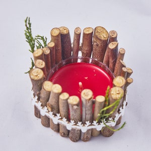 Puede incluir: Una vela roja se encuentra dentro de un portavelas rústico de ramas. El portavelas está decorado con encaje blanco y pequeñas ramitas de vegetación.