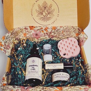 May include: A cardboard gift box containing a 4 oz bottle of Alquimia de raiz hair oil, a small roller bottle, a round jar, and a pink silicone scalp massager. The box interior features a floral pattern and the text Apotecario Moderno by Clary.