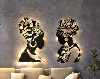 Könnte beinhalten: Zwei Wandkunst-Silhouetten aus schwarzem Metall, die Profile von Personen mit Kopftüchern darstellen. Die ausgeschnittenen Designs werden von hinten durch warmes LED-Licht beleuchtet, was einen Leuchteffekt um die dunklen Metallfiguren erzeugt.