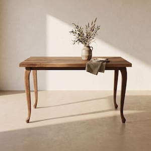 May include: A rectangular wooden table with four curved legs. A ceramic vase containing dried branches sits on the table surface, accompanied by a folded beige cloth napkin draped over the edge of the tabletop.