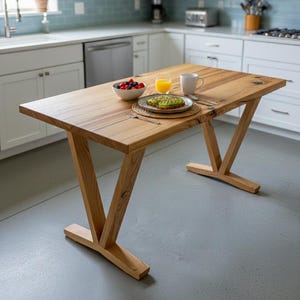 May include: A light-colored wooden kitchen table with a rectangular top and V-shaped legs. The table is set with a bowl of berries, a plate of avocado toast, a glass of orange juice, and a mug. The kitchen has white cabinets and a blue tile backsplash.