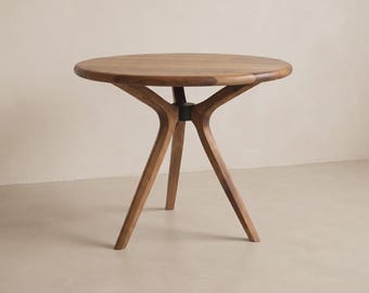 Op de afbeelding: Een ronde houten tafel met een glad, cirkelvormig tafelblad met een zichtbare natuurlijke houtnerf. De tafel wordt ondersteund door drie schuine, taps toelopende houten poten die samenkomen in een donkere centrale naaf onder het blad.