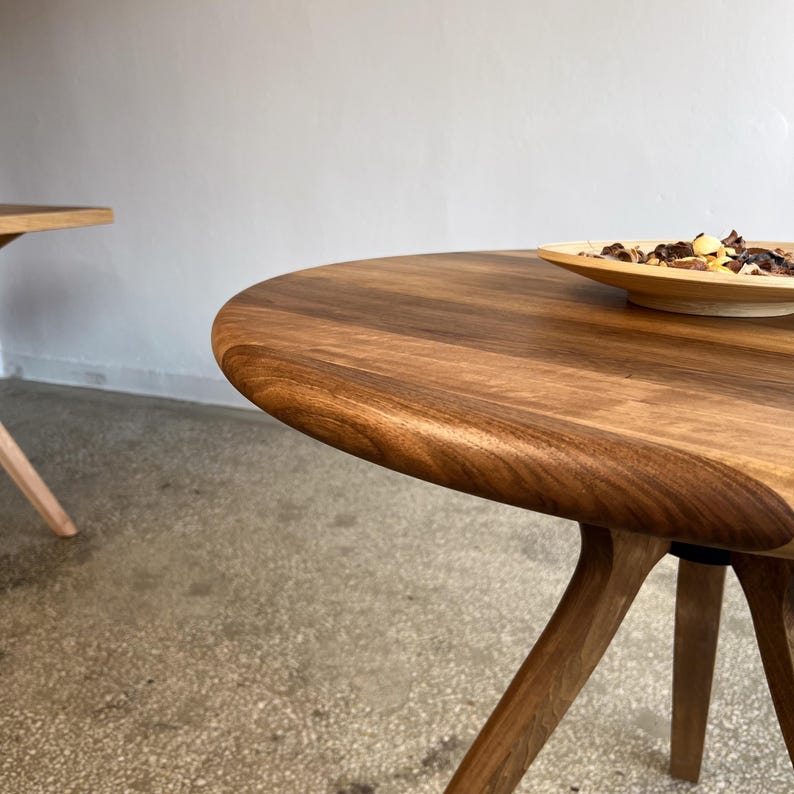 Angled close-up view of round solid walnut dining table highlighting smooth curved edge, flowing natural wood grain patterns, and hand-finished surface, sculptural base connection subtly visible beneath tabletop, warm walnut tones enhanced by soft natural lighting, partial view of decorative bowl adding scale and context, minimal interior setting emphasizing material quality and craftsmanship, ideal for modern dining spaces, durable hardwood construction with custom size options available