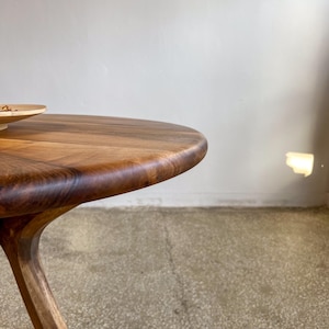 Close-up side detail of round solid walnut dining table highlighting smooth curved edge profile, rich natural wood grain, and hand-finished surface, sculpted transition from tabletop to base showcasing fine craftsmanship, warm dark walnut tones enhanced by natural light reflections, minimal composition focusing on material quality and form, durable hardwood construction ideal for modern dining interiors, custom size options available