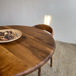 Angled top view of round solid walnut dining table highlighting rich natural wood grain patterns, smooth handcrafted surface, and softly rounded edge profile, warm walnut tones enhanced by natural light reflections, minimal styling with decorative bowl placed on tabletop, partial view of chair in background adding context without distracting from product, ideal for kitchen, dining room, or small space interiors, durable hardwood construction with custom size options available