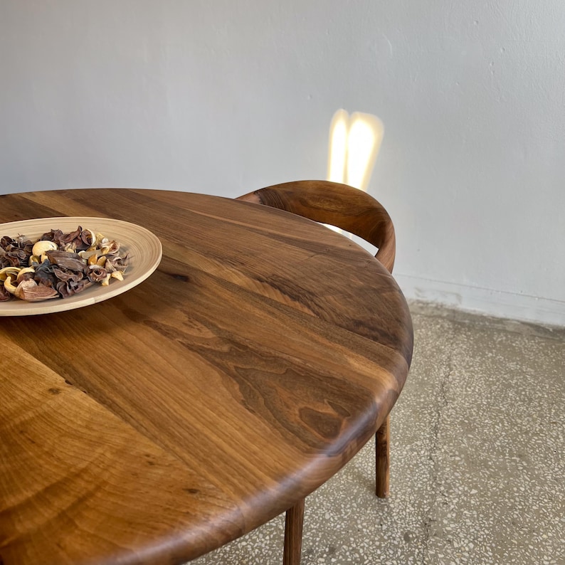 Angled top view of round solid walnut dining table highlighting rich natural wood grain patterns, smooth handcrafted surface, and softly rounded edge profile, warm walnut tones enhanced by natural light reflections, minimal styling with decorative bowl placed on tabletop, partial view of chair in background adding context without distracting from product, ideal for kitchen, dining room, or small space interiors, durable hardwood construction with custom size options available