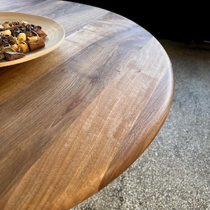 Close-up angled detail of round solid walnut dining table highlighting flowing natural wood grain transitions, tonal variations, and smooth hand-finished surface, softly rounded edge profile with organic shaping, warm walnut hues enhanced by directional natural light, partial view of decorative bowl adding scale and context, minimal composition emphasizing material quality, texture, and craftsmanship, ideal for modern dining interiors, durable hardwood construction with custom sizes