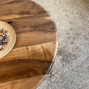 Close-up detail of round solid walnut dining table surface highlighting rich natural wood grain patterns, tonal variations, and organic texture transitions, smooth handcrafted finish with soft curved edge detail, warm brown hues creating a refined and natural aesthetic, partial view of decorative bowl on tabletop, high quality hardwood craftsmanship emphasizing unique character of walnut wood, suitable for modern dining interiors with custom size options available
