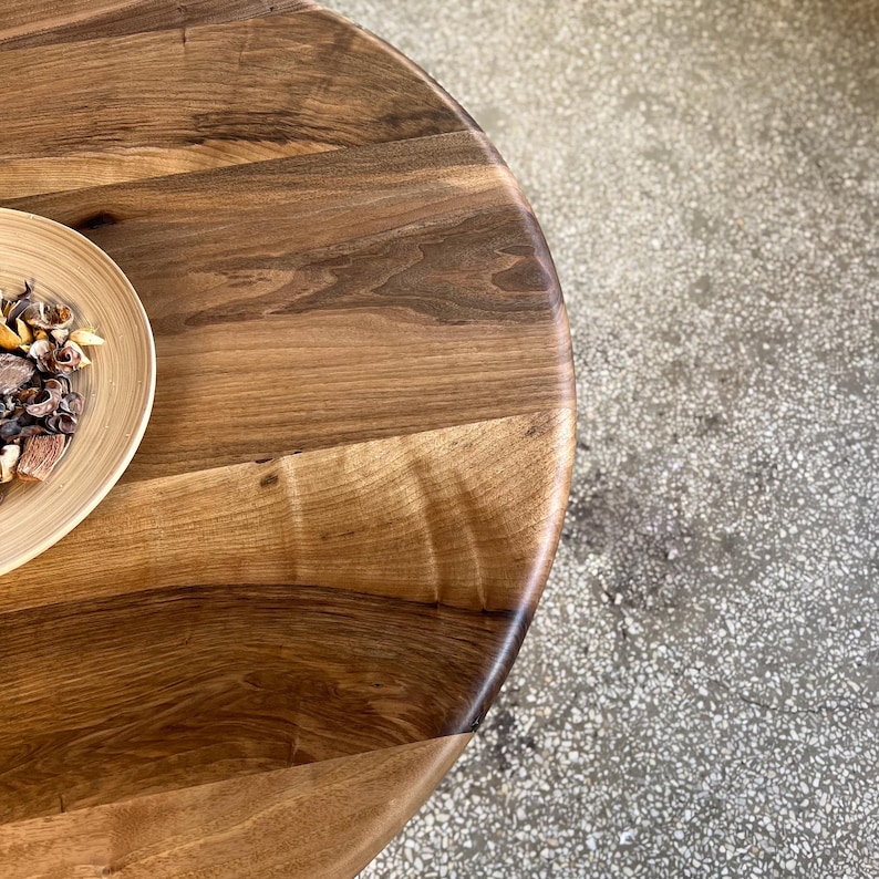 Close-up detail of round solid walnut dining table surface highlighting rich natural wood grain patterns, tonal variations, and organic texture transitions, smooth handcrafted finish with soft curved edge detail, warm brown hues creating a refined and natural aesthetic, partial view of decorative bowl on tabletop, high quality hardwood craftsmanship emphasizing unique character of walnut wood, suitable for modern dining interiors with custom size options available