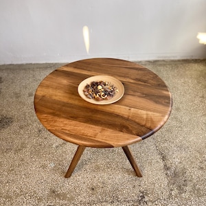High angle view of round solid walnut dining table showcasing rich natural wood grain variations, warm brown tones, and smooth handcrafted finish, circular tabletop with organic flowing patterns and soft edge detailing, minimal styling with decorative bowl placed at center, sculptural tripod base subtly visible, natural light reflections adding depth and warmth, suitable for kitchen, dining room, or small space interiors, durable hardwood construction with custom size options available