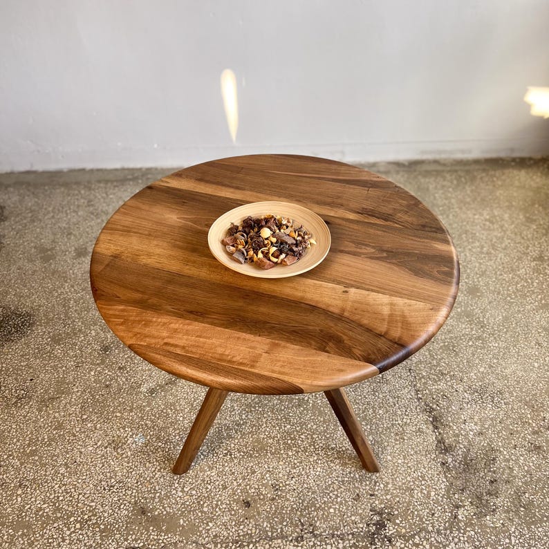 High angle view of round solid walnut dining table showcasing rich natural wood grain variations, warm brown tones, and smooth handcrafted finish, circular tabletop with organic flowing patterns and soft edge detailing, minimal styling with decorative bowl placed at center, sculptural tripod base subtly visible, natural light reflections adding depth and warmth, suitable for kitchen, dining room, or small space interiors, durable hardwood construction with custom size options available