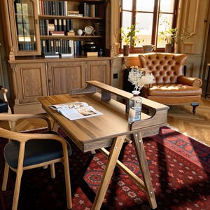 May include: A wooden desk with a built-in shelf and angled legs sits on a red patterned rug. A chair with a black seat and wooden frame is to the left. A leather armchair and a wooden bookcase are in the background.