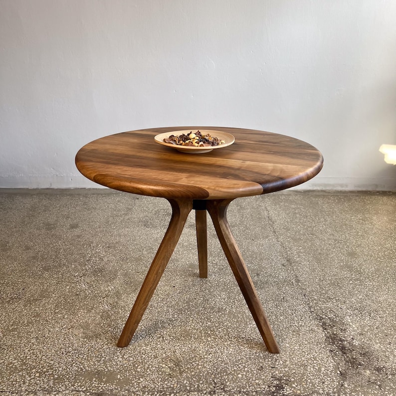 Round solid walnut dining table with smooth circular tabletop and rich natural wood grain, handcrafted wooden table featuring a sculptural tripod style base with angled legs, warm dark walnut tones and organic edge details creating a refined and modern look, minimal setup with decorative bowl on top, ideal for kitchen, dining area, or small space interiors, durable hardwood construction with timeless design and custom size options available