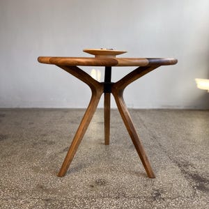 Side profile view of round solid walnut dining table highlighting sculptural tripod base design with angled legs and central support connection, smooth circular tabletop with rich natural wood grain and hand-finished surface, balanced and symmetrical structure emphasizing craftsmanship and stability, warm walnut tones enhanced by soft natural lighting, minimal setting with decorative bowl on top, ideal for modern dining interiors, durable hardwood construction with custom size options available