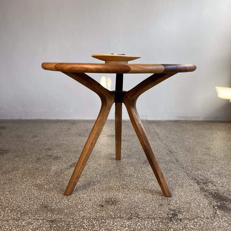 Side profile view of round solid walnut dining table highlighting sculptural tripod base design with angled legs and central support connection, smooth circular tabletop with rich natural wood grain and hand-finished surface, balanced and symmetrical structure emphasizing craftsmanship and stability, warm walnut tones enhanced by soft natural lighting, minimal setting with decorative bowl on top, ideal for modern dining interiors, durable hardwood construction with custom size options available