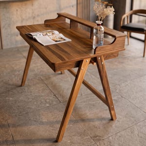 Op de afbeelding: Een houten bureau met een donkerbruine nerf, voorzien van een A-vormig onderstel, een verhoogde achterplank en een lade. Een open boek ligt plat op het oppervlak naast een doorzichtige glazen fles met gedroogde bloemen.