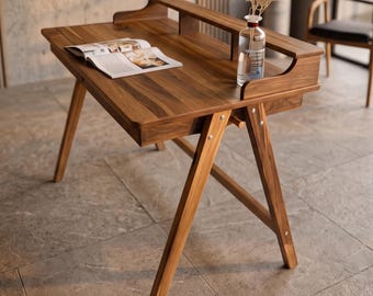 May include: A wooden desk with a dark brown grain, featuring an A-frame leg structure, a raised rear shelf, and a single drawer. An open book lies flat on the surface next to a clear glass bottle containing dried flowers.