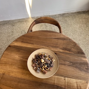Top view of round solid walnut dining table highlighting natural wood grain patterns, warm dark walnut tones, and smooth handcrafted surface, circular tabletop with subtle organic variations and refined edge detail, minimal styling with decorative bowl placed at center, sculptural wooden base partially visible, suitable for kitchen, dining room, or small space interiors, durable hardwood construction with timeless design and custom size options available